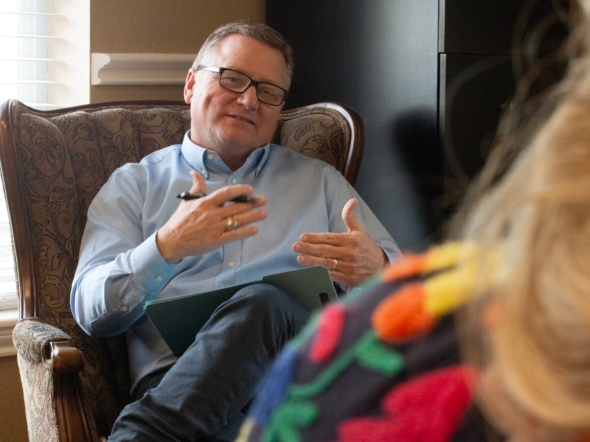 A man in a light blue button-up shirt seated in a patterned chair, gesturing with his hands while speaking to someone off camera