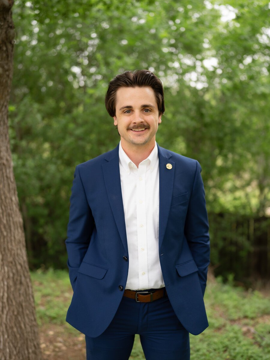 Portrait of Turner Howk in a navy blazer and white shirt outdoors with green foliage